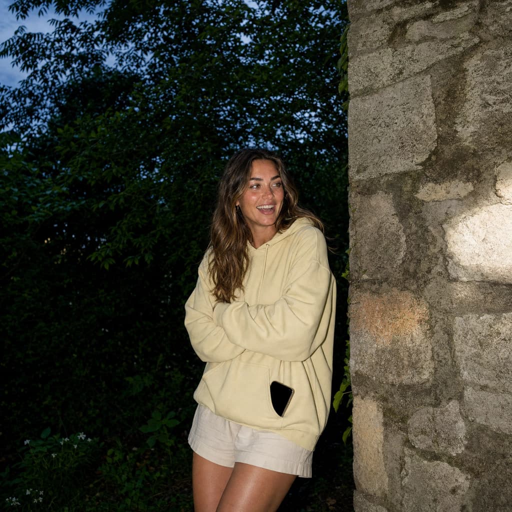 A woman in a butter-yellow hoodie laughing against a stone wall, arms folded and her phone tucked into a pocket.