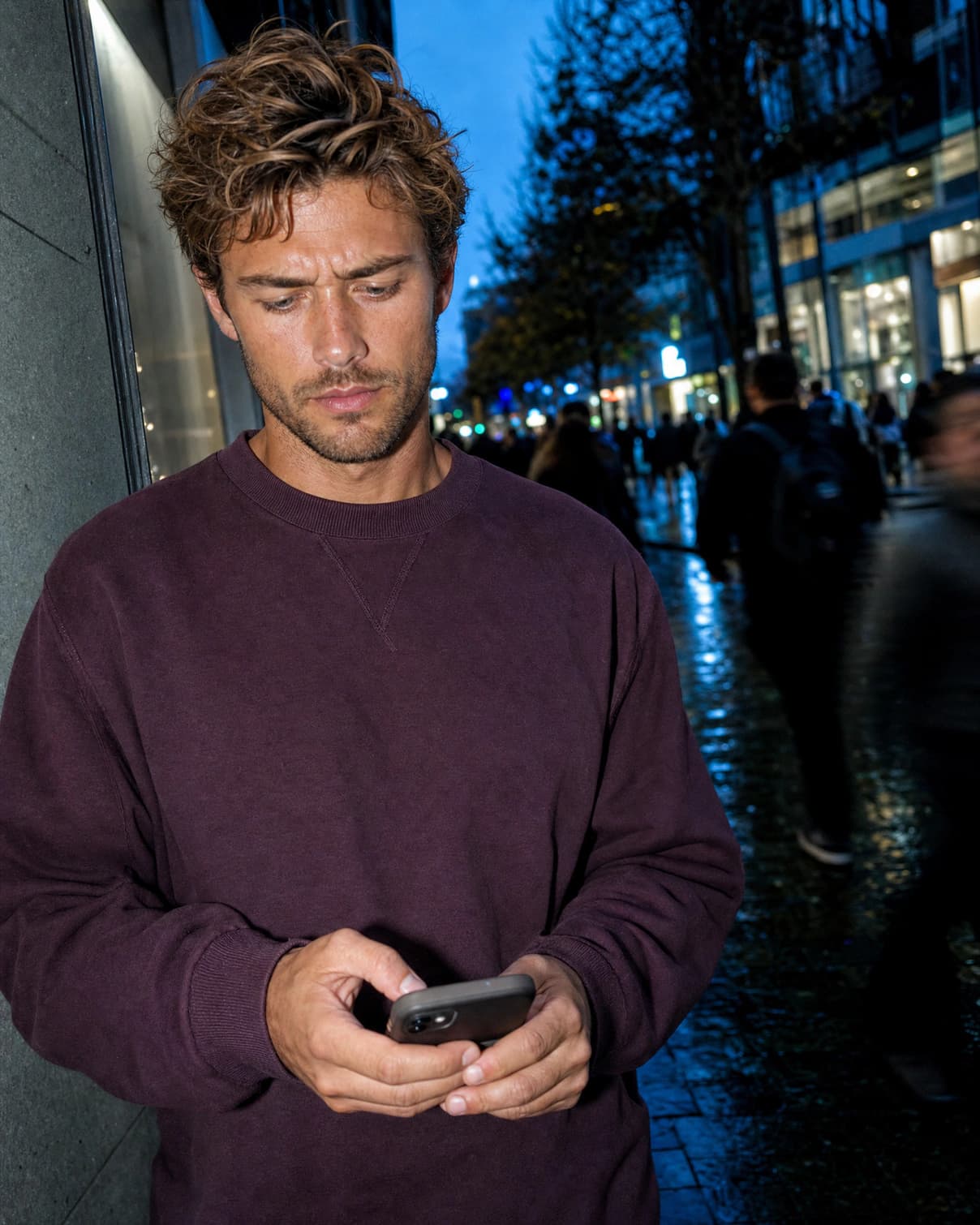 A man in a plum sweatshirt on a busy street at dusk, looking down at his phone with a furrowed brow.