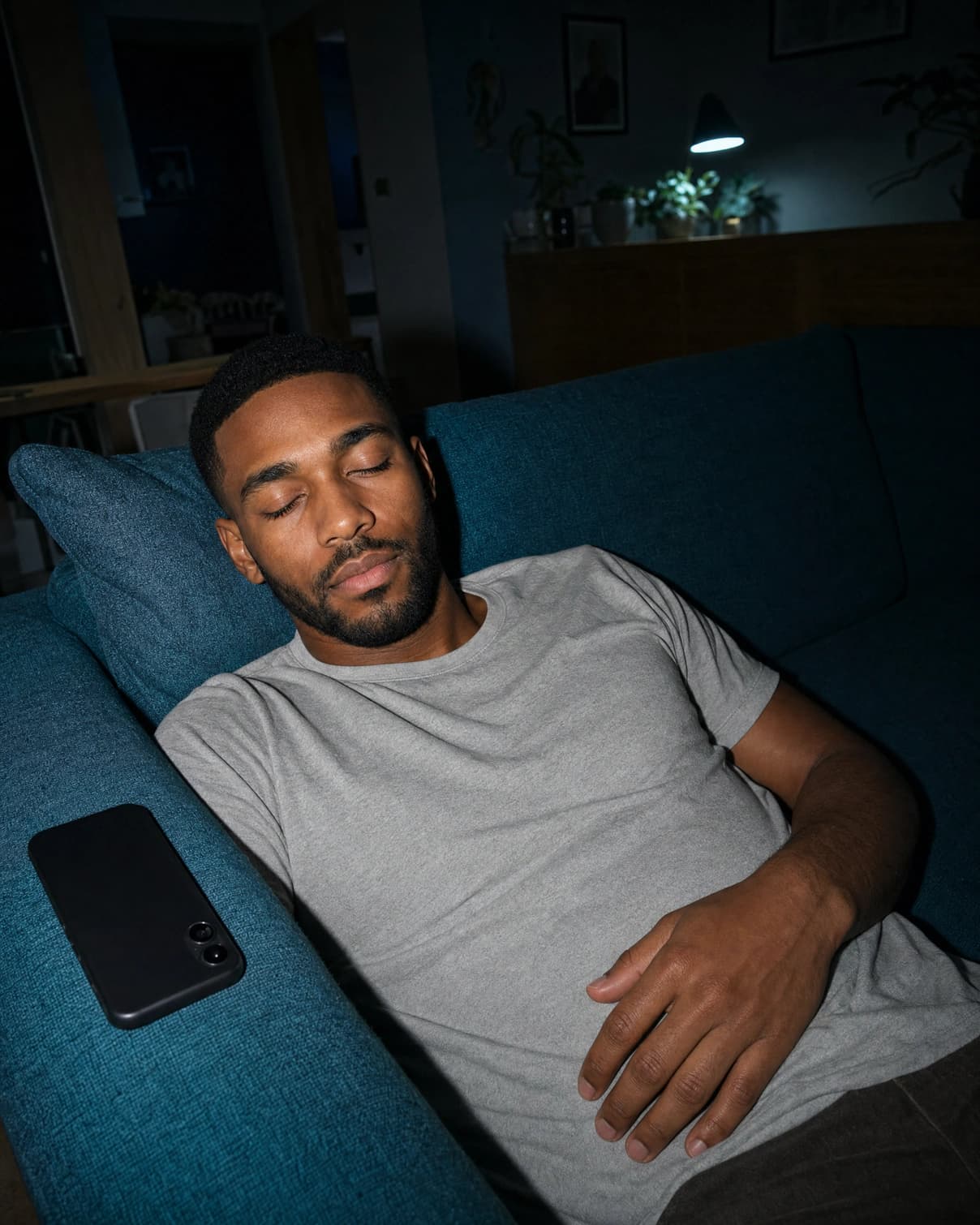 A man asleep on a sofa in a dark living room, with his phone face down on the arm beside him.