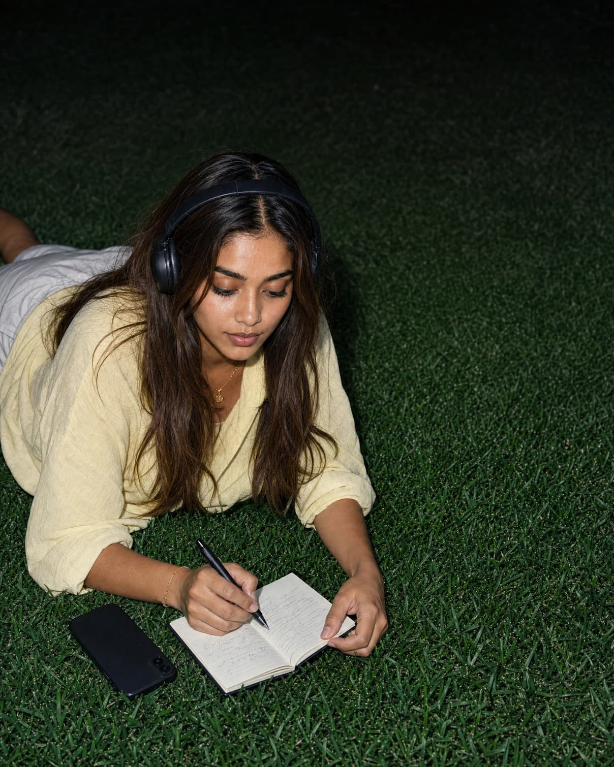 A woman in a yellow top lying on grass at night, wearing headphones and writing in a notebook, with her phone face down beside her.