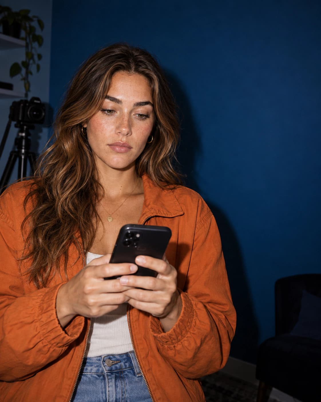A woman in an orange jacket looking at her phone in a home studio, with a camera on a tripod behind her.