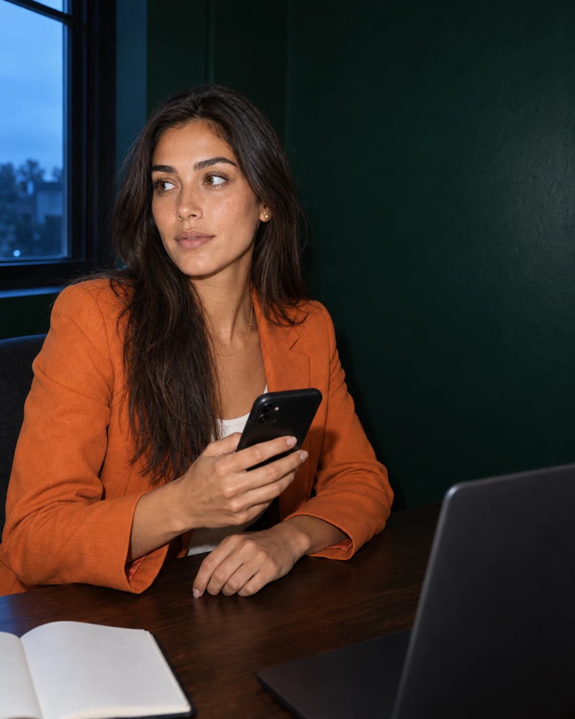 A woman in an orange blazer at a desk with her phone, notebook and laptop.