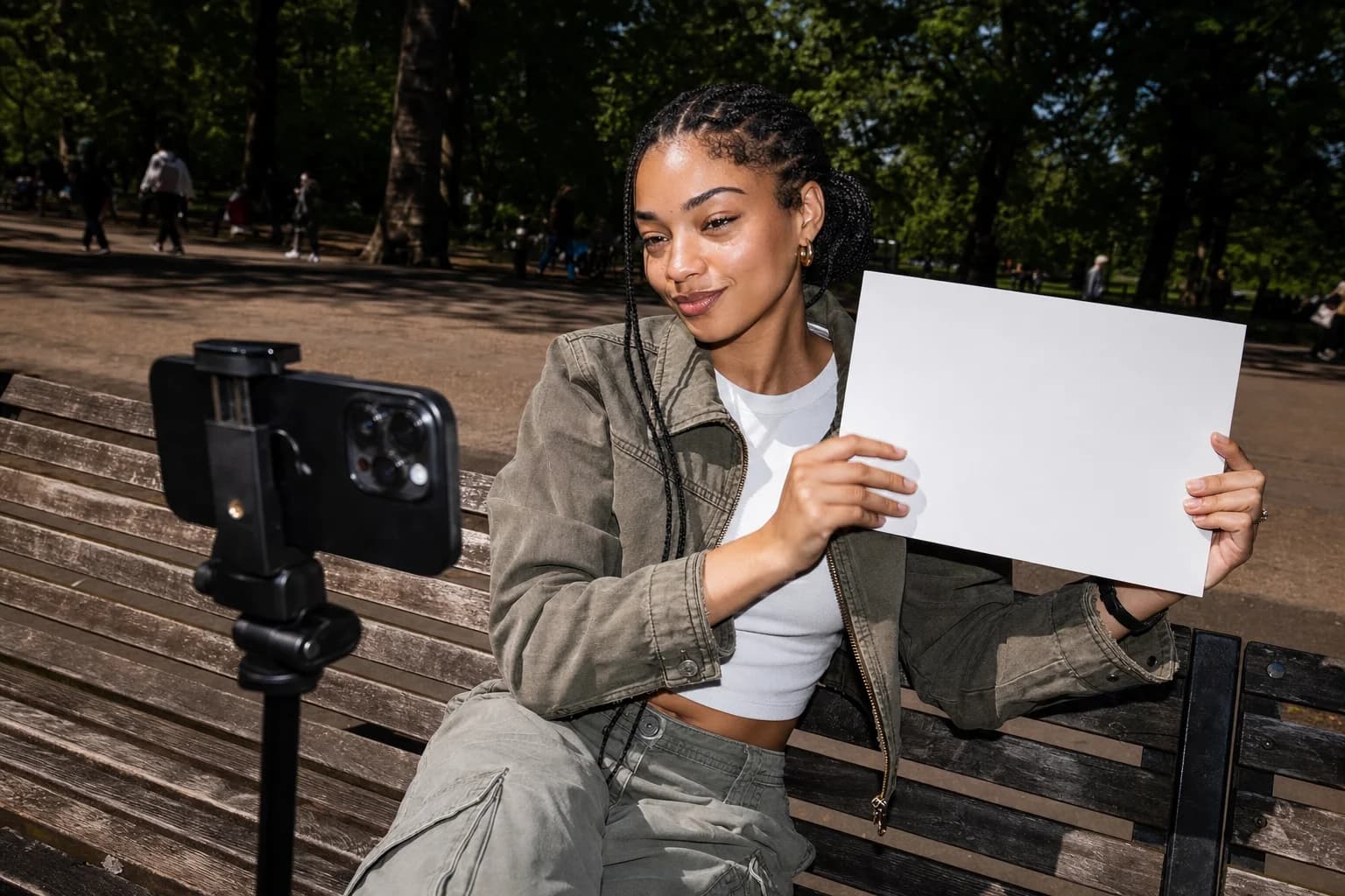 Young woman on a sunlit park bench holds a blank white card up to a phone on a mini tripod, smirking at the lens.