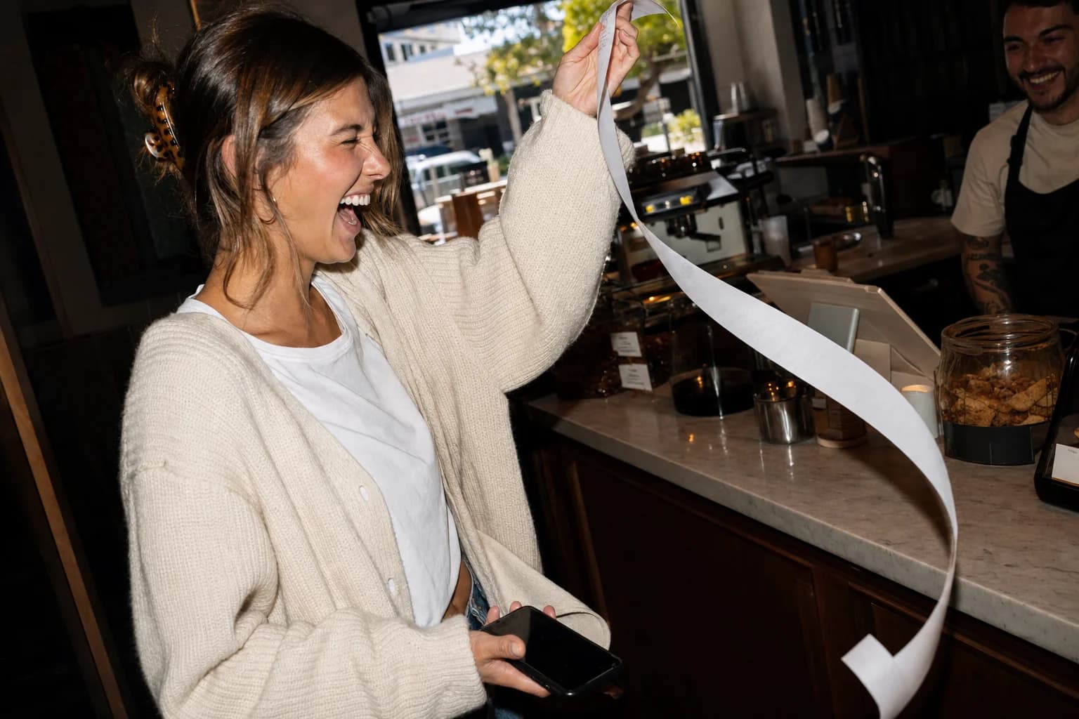 Young woman in a cream cardigan laughs at a long till receipt she holds up at a sunlit café counter, phone in her other hand.
