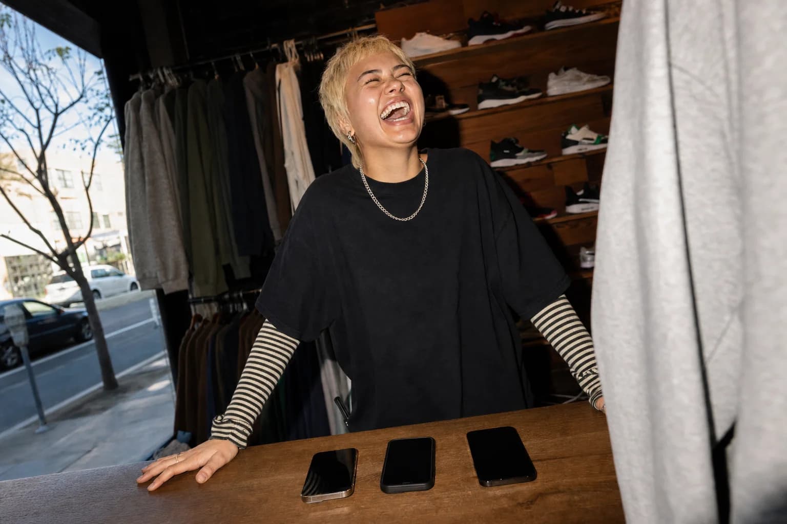 Young woman behind a streetwear shop counter laughs at three phones lined up in front of her, storefront window bright behind.