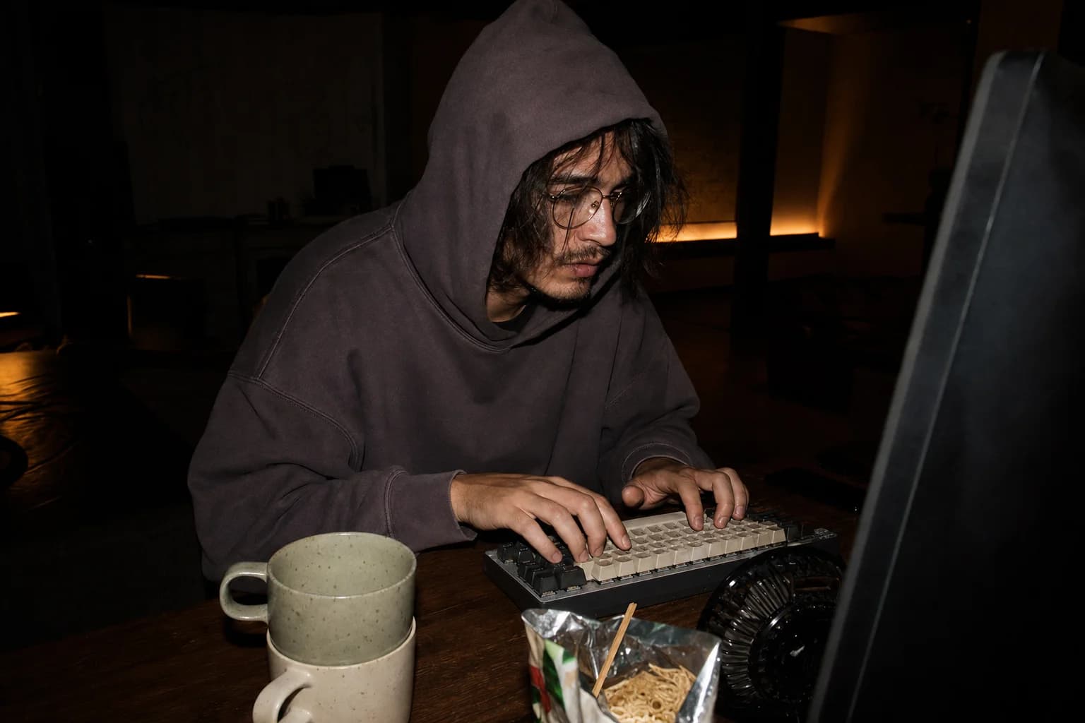 Hooded man in glasses types on a mechanical keyboard late at night, beside stacked mugs and instant noodles.