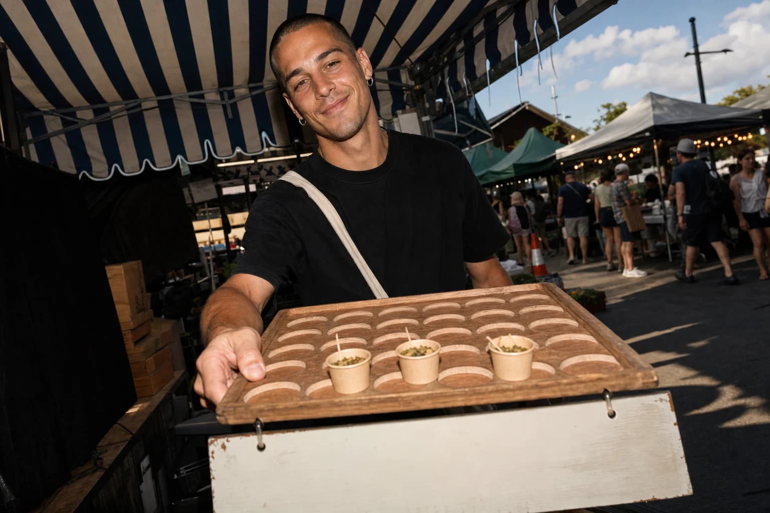 Young man under a striped market canopy holds out a wooden sample tray with only three cups left, shoppers blurred behind him.