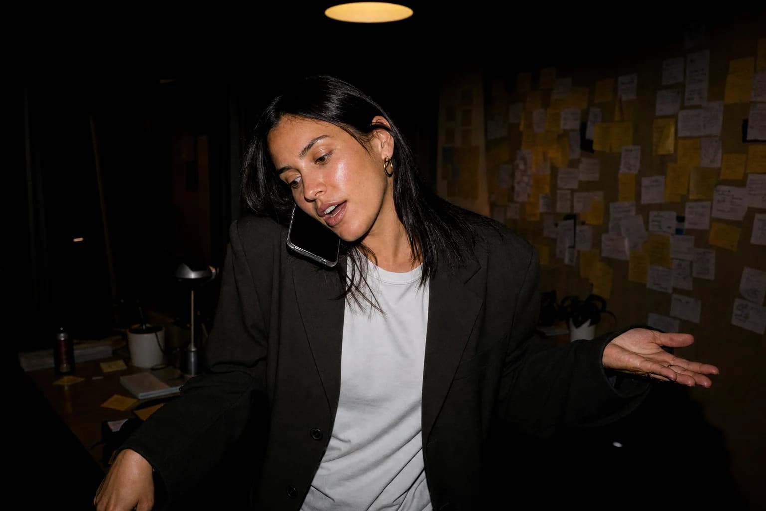 Woman in an oversized blazer takes a call in a dark office, mid-gesture, a wall of sticky notes behind her.
