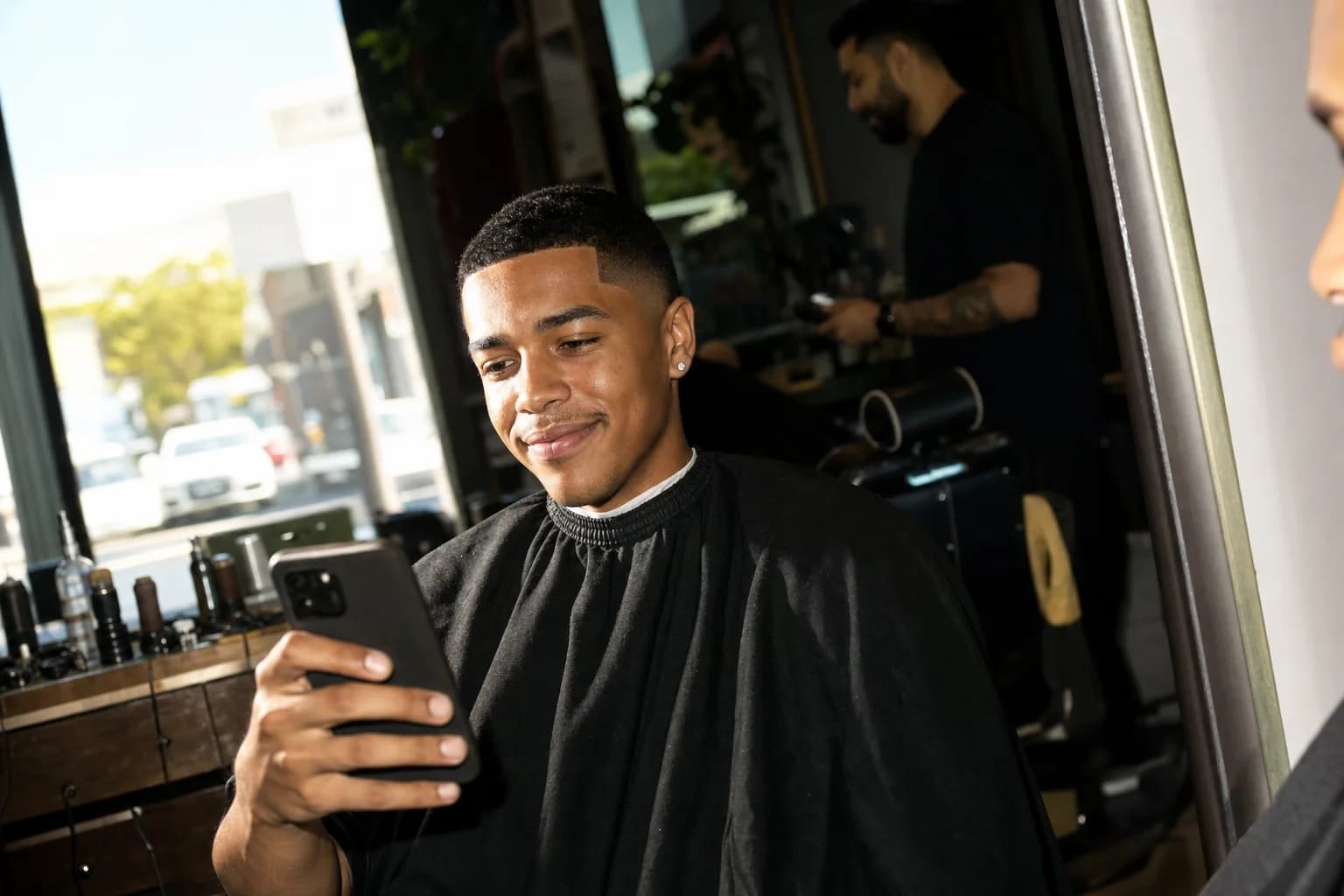 Young man in a barber's cape checks his phone with a content smirk in a sunlit barbershop chair, barber blurred behind.