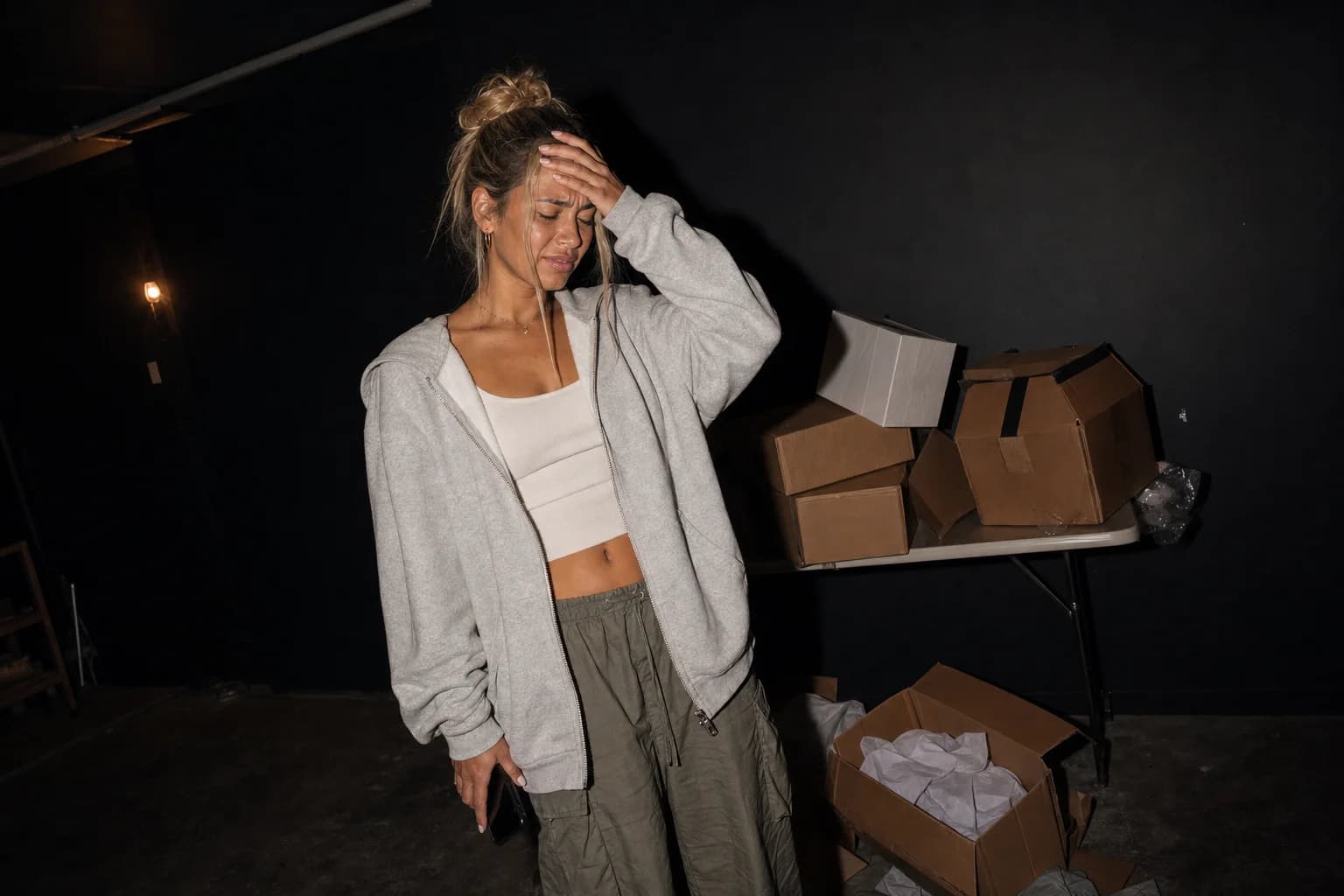 A woman winces with a hand to her forehead in a dim studio, phone at her side, toppled boxes on the table behind her.