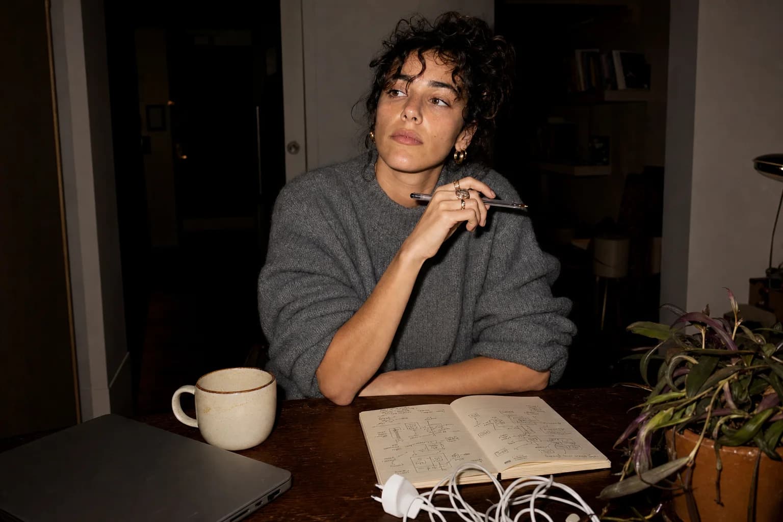 Woman in a grey knit pauses at a night desk, pen to her chin, over a notebook of hand-drawn boxes and arrows.