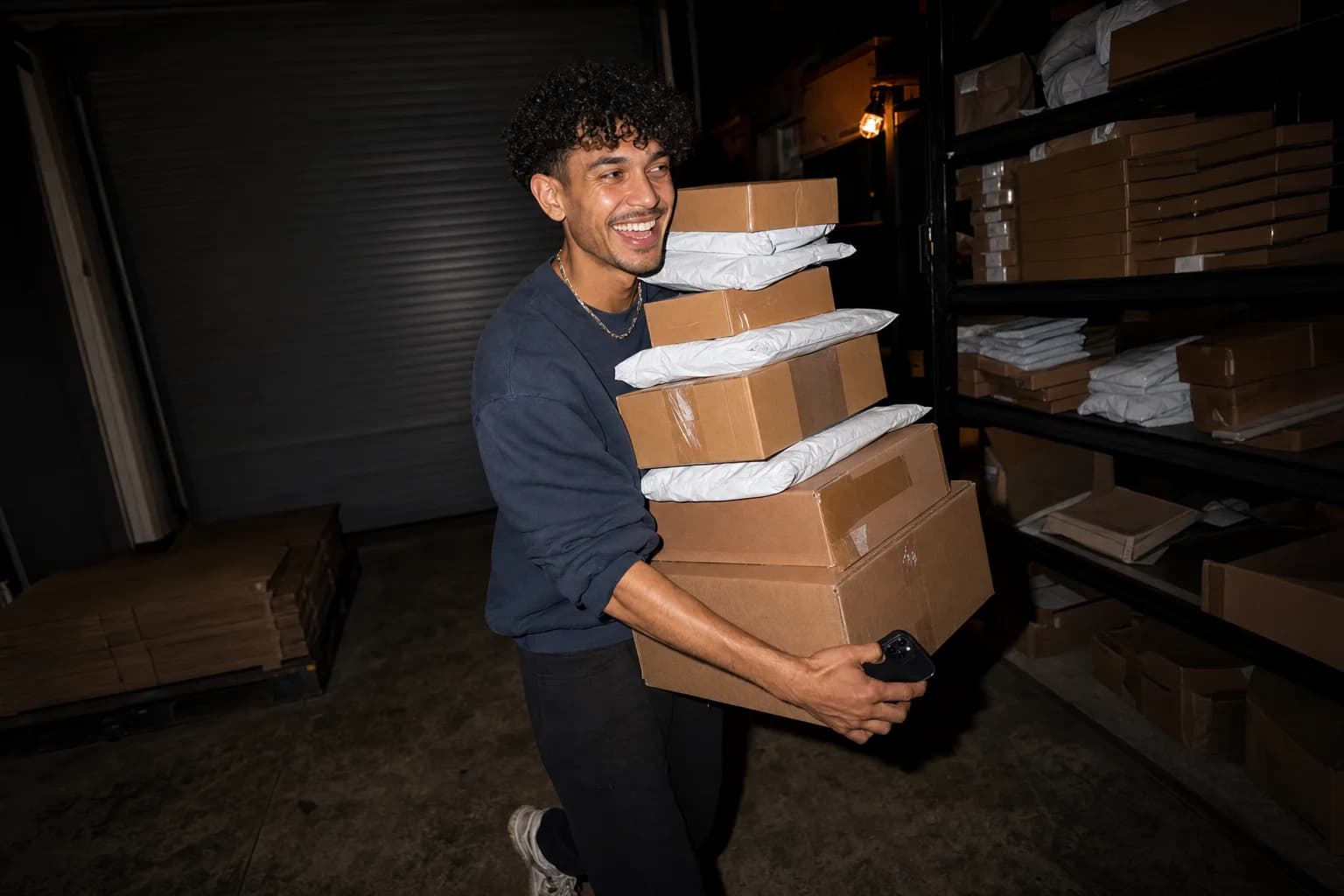 A man grins mid-stride through a dark warehouse, carrying a tall stack of parcels with his phone in one hand.