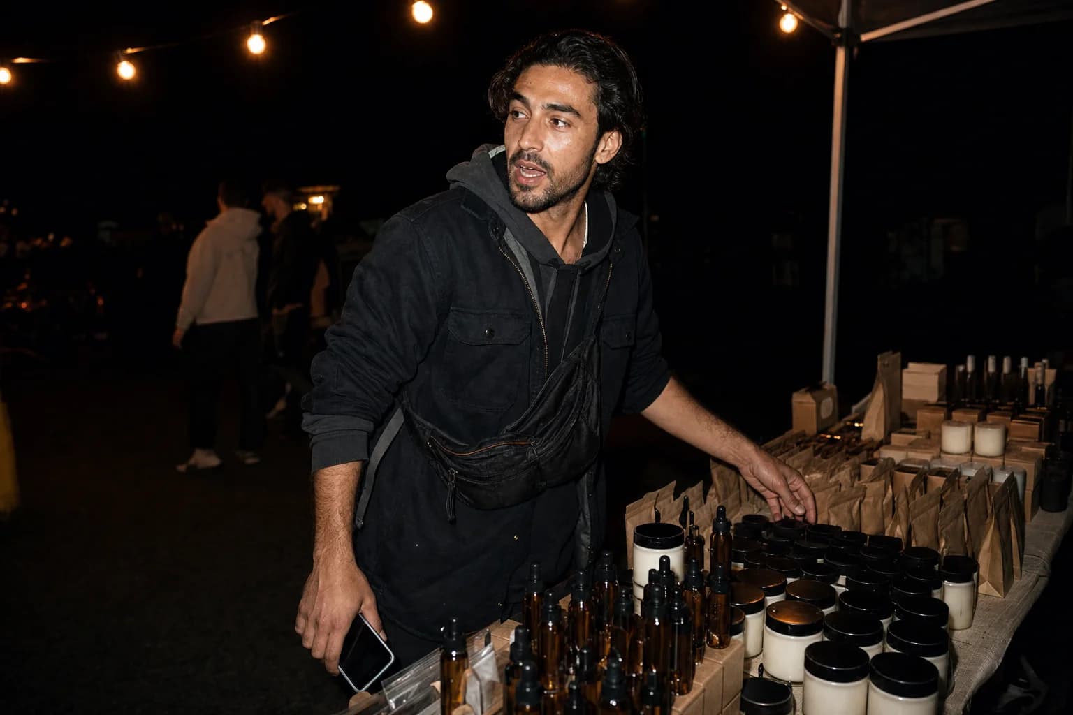 A man works a night market stall of candles and amber bottles, phone in one hand, turning to talk to someone off-frame.