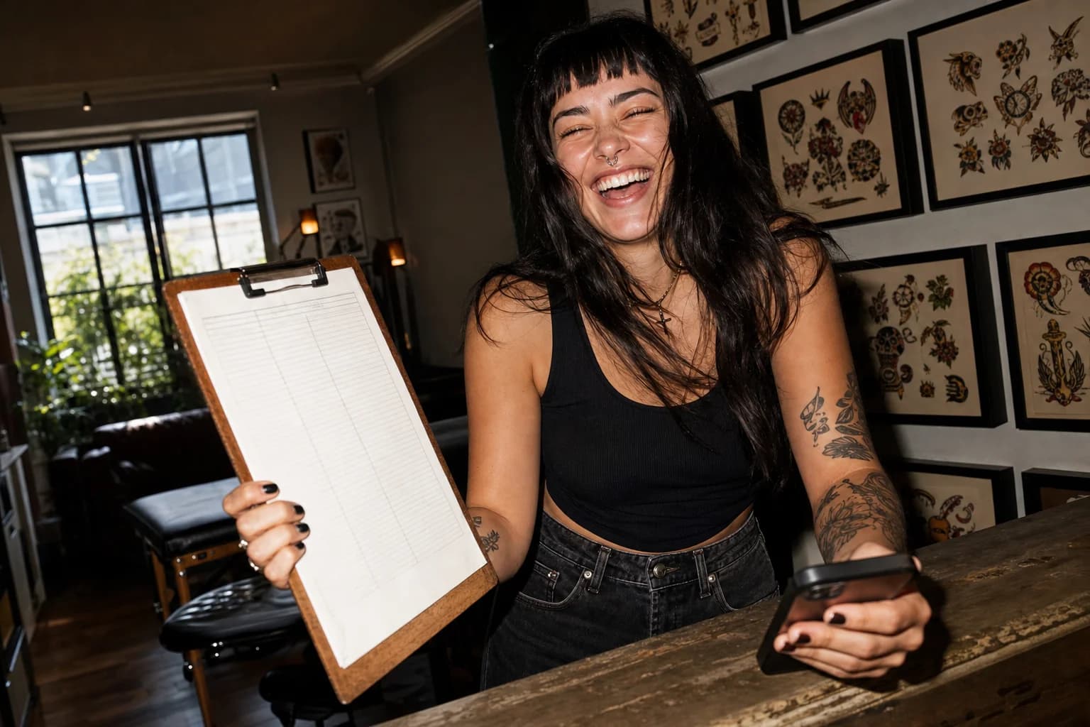 Young woman at a sunlit tattoo-studio front desk laughs while holding up a paper sign-in clipboard next to her phone.