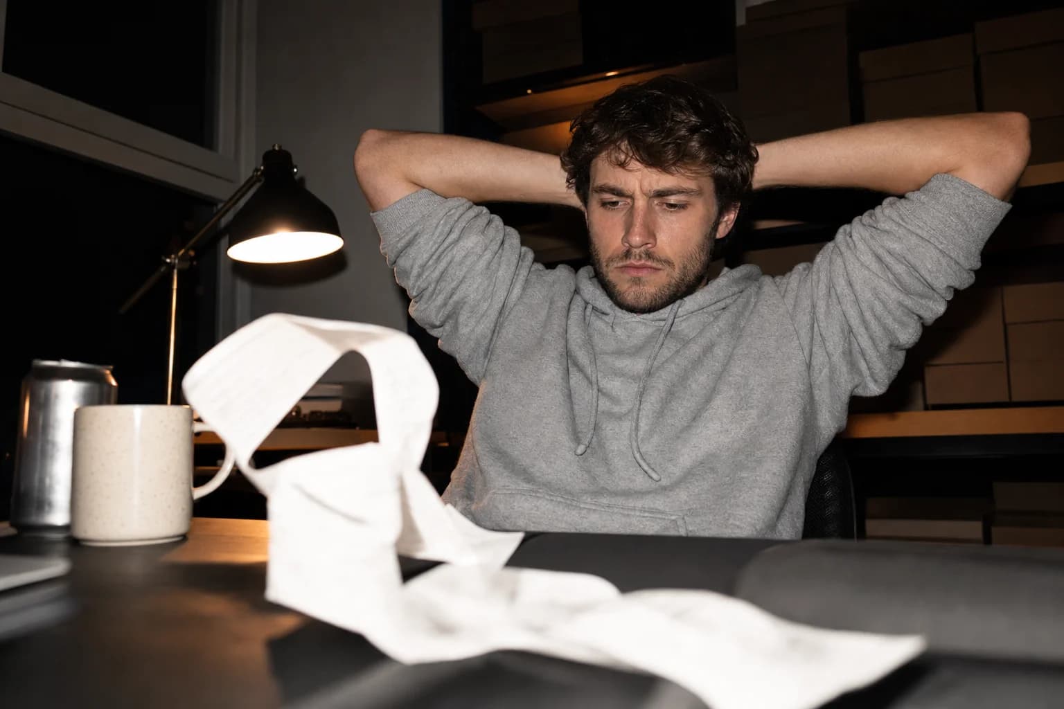 A man leans back at a dark desk, hands behind his head, frowning at a long paper receipt tape spilling onto the floor.