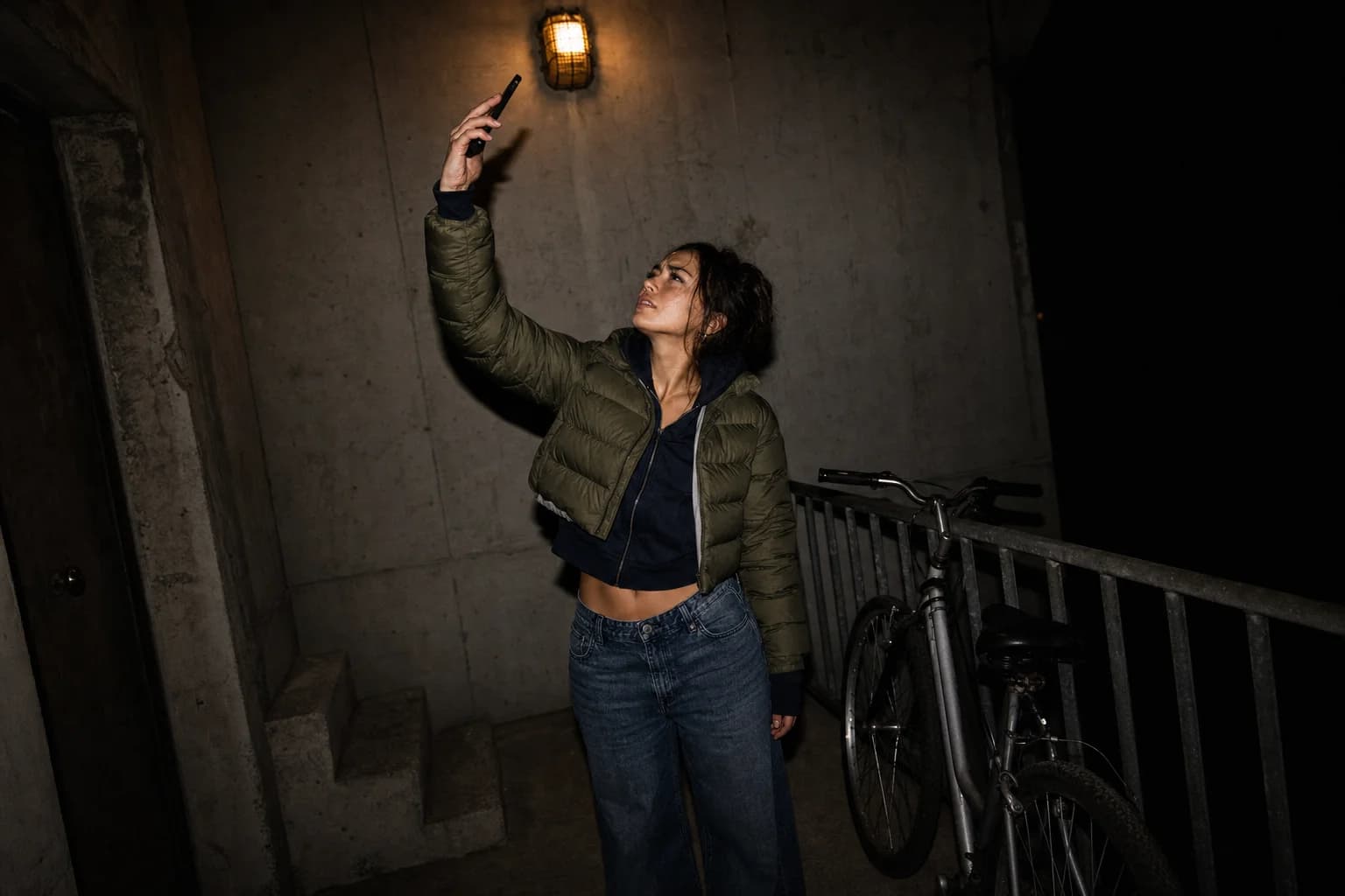 A woman in a dim concrete stairwell holds her phone up at arm's length, squinting at it, hunting for a signal.