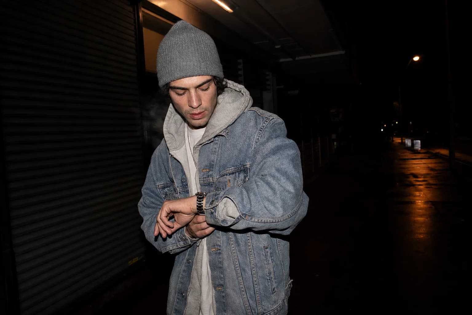 Man in a grey beanie and denim jacket checking his watch outside a shuttered shopfront on a dark street