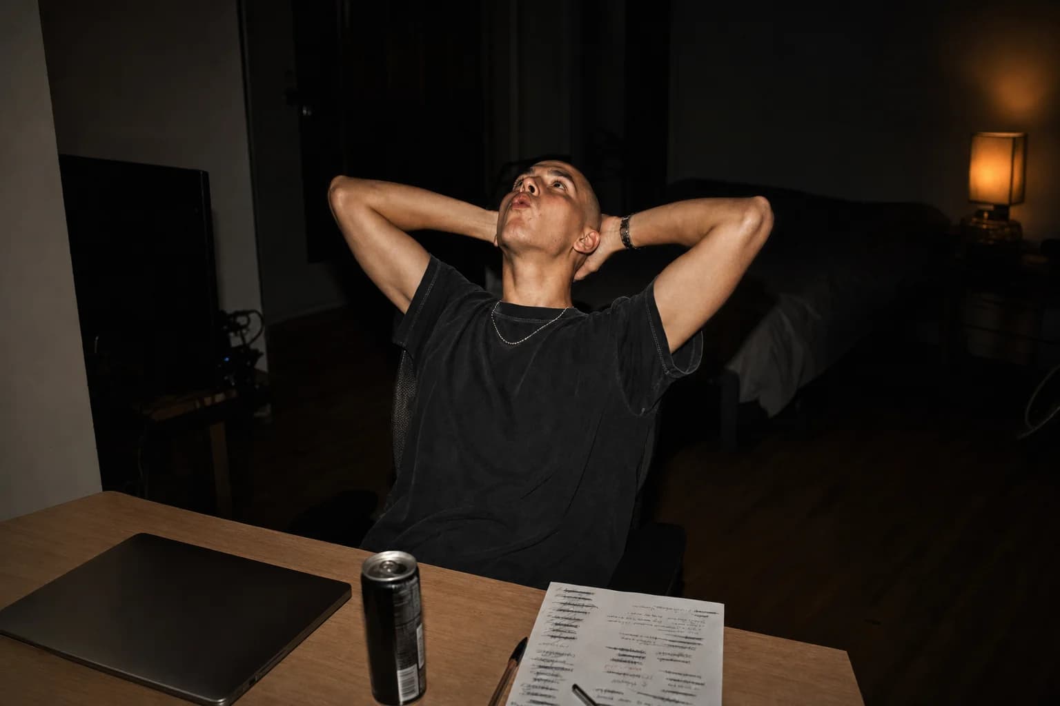 Man in a black tee leans back from his desk at night, hands behind his head, staring up at the ceiling.